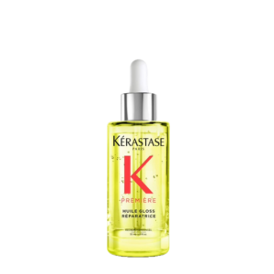 A clear bottle of Kérastase Première Huile Gloss Réparatrice hair oil with a white dropper cap, filled with yellow liquid, is shown against a plain white background. The elegant packaging highlights the luxurious blend of Première Huile and Reparatrice ingredients designed to restore shine and vitality to your hair. | Blush Salon Collective - Fargo, ND